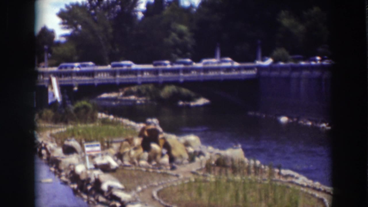 Cars crossing a bridge over a river with rocks, grass, and trees nearby.