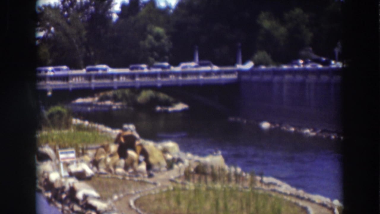Cars cross the Truckee river bridge in a soft 8mm frame, with stones, grasses, water, and trees visible around the span.