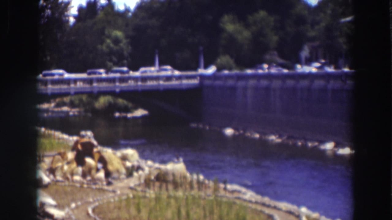 Truckee bridge traffic continues above the river, framed by foreground stones, grasses, tree shade, and the soft edges of 8mm film.