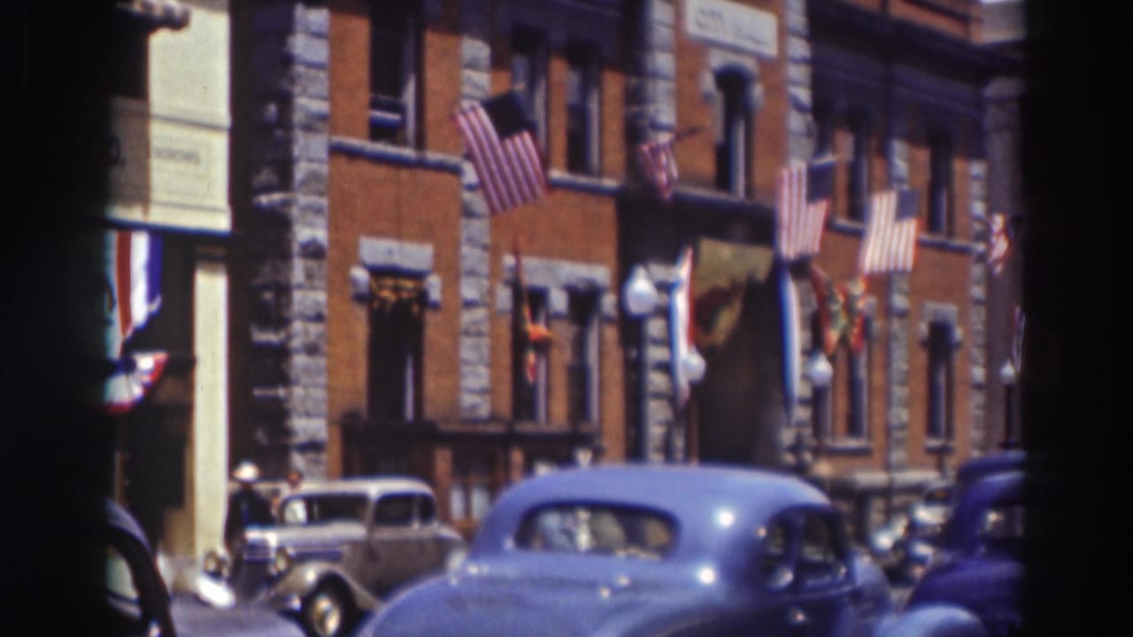 Brick street building with American flags and parked 1930s cars.