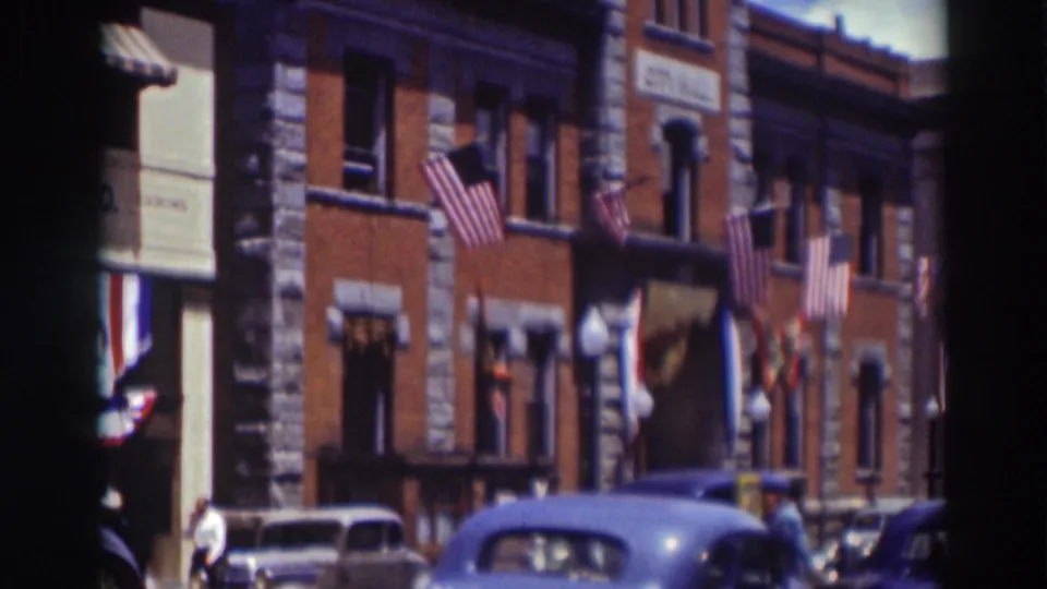 1937 Truckee footage frames a decorated brick facade with American flags, hanging banners, and period cars at street level.