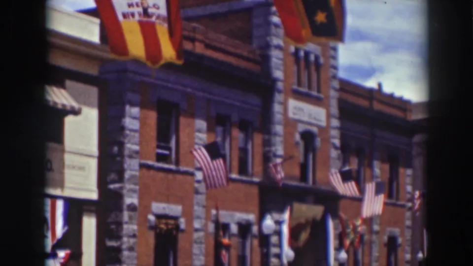 Late-frame view of the 1937 Truckee street scene with flags, banners, a brick-and-stone facade, and parked period automobiles.