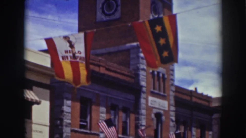 1937 Truckee home-movie frame showing a red-and-yellow banner, a vertical star banner, American flags, and brick storefront architecture.
