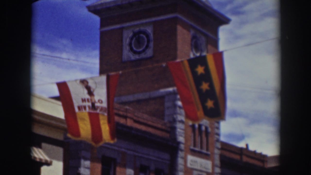 Brick clock tower with two hanging flags, power lines, and blue sky.