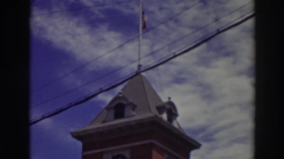 Truckee 1937 home-movie frame of a clock tower roof and flagpole against a blue, clouded sky with utility lines crossing.