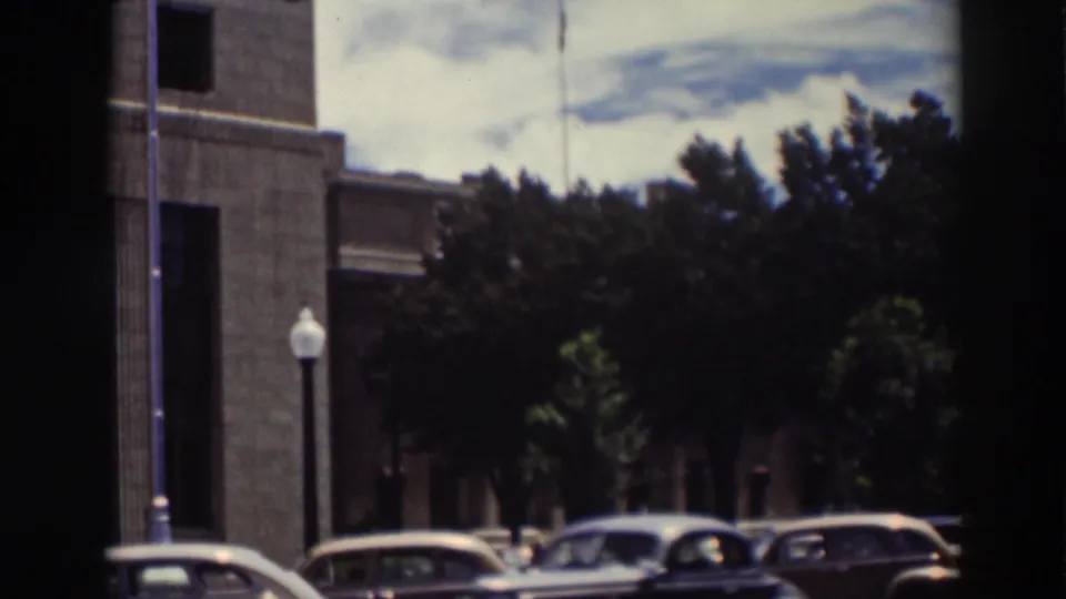 Truckee 1937 8mm frame showing a civic building edge, trees, streetlamp, and several parked early automobiles along the street.