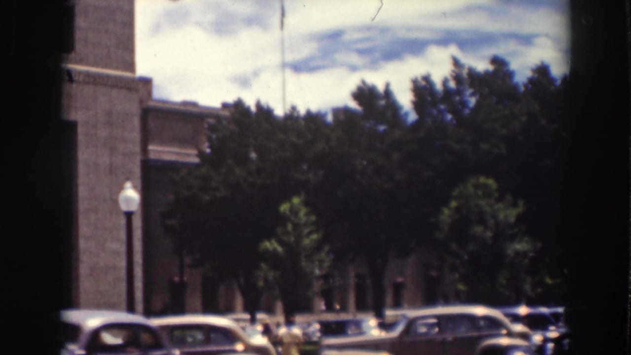Parked 1930s cars along a tree-lined street beside a large brick building.