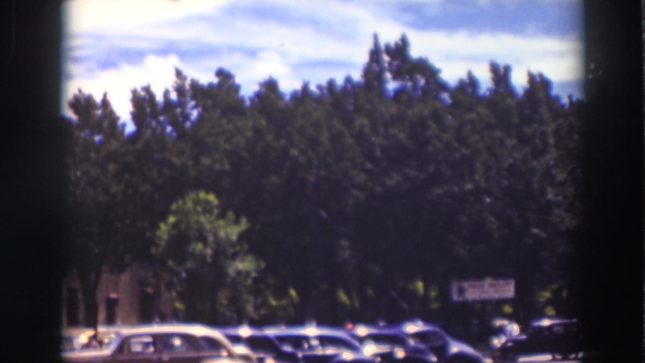 1937 Truckee home-movie frame showing tall trees, roadside cars, and a partly cloudy sky from a moving viewpoint.