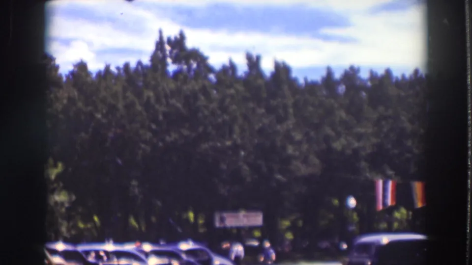 Soft 8mm frame of Truckee roadside trees, cars, small distant pedestrians, and a vertical banner or flag at frame right.
