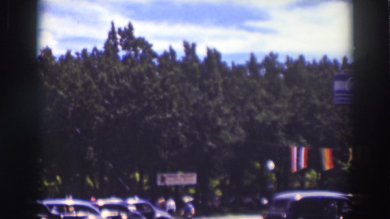 Tree-lined street with period cars, striped banners, and a partial roadside sign.
