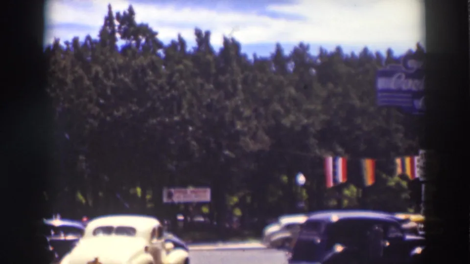 Late-frame Truckee street view with prewar cars crossing the foreground, dense trees, striped banners, and partial signage.