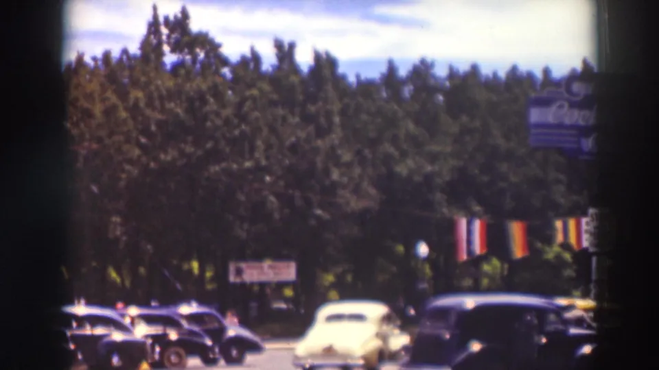 1937 Truckee street scene showing a pale yellow automobile among dark 1930s cars, flags, trees, and a distant sign.