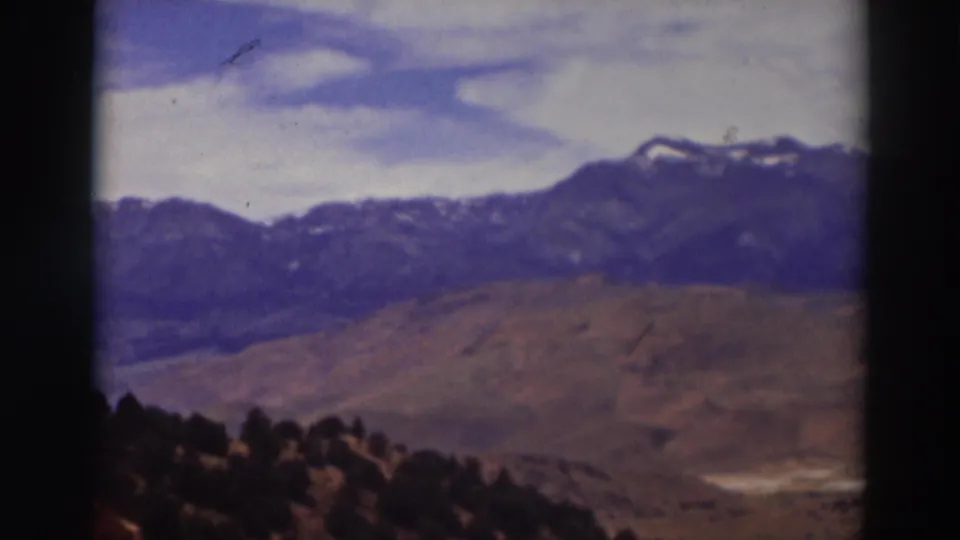 1937 8mm view near Truckee, California, showing distant snow-dusted mountains, arid hills, clouds, and dark foreground trees.