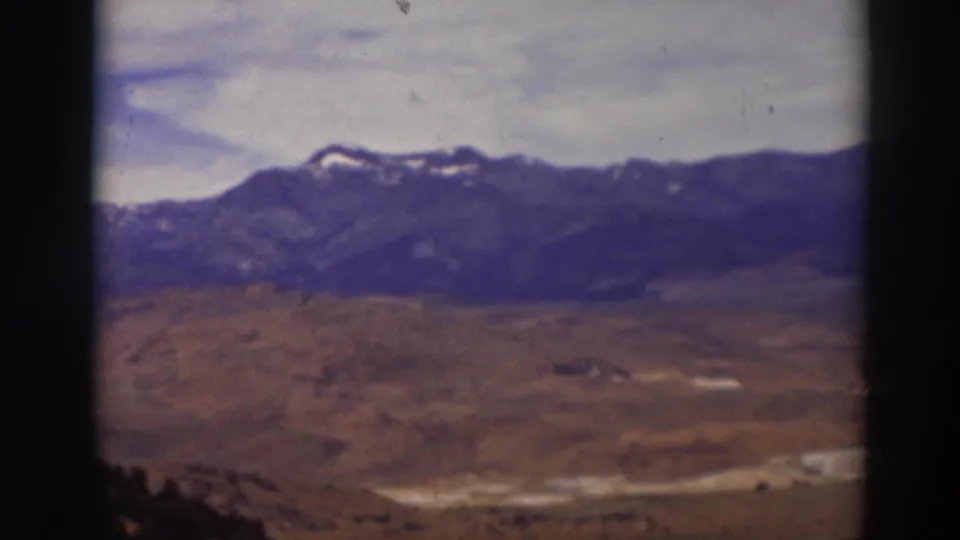 Late-frame 8mm view near Truckee with distant snow-dusted mountains, dry hills, pale clouds, and dark foreground trees.