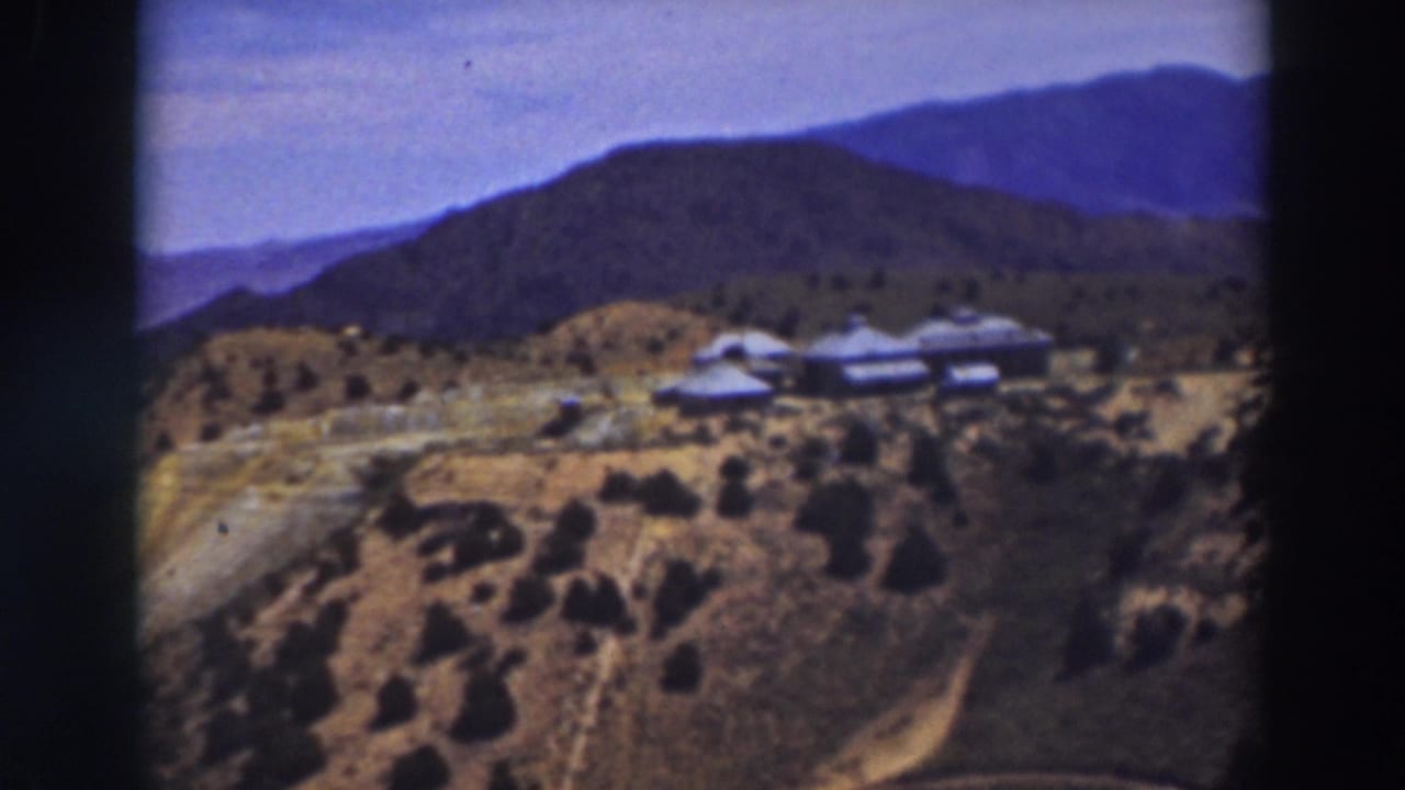 1937 Truckee-area 8mm frame showing a hillside building cluster, scattered trees, dry slopes, and distant blue mountains.