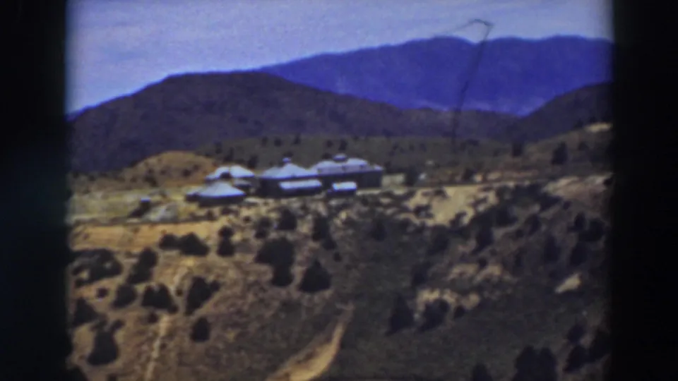 1937 8mm view near Truckee showing pale-roofed structures, uneven hillside terrain, scattered trees, and blue mountain backdrop.
