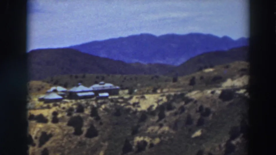 Late-1930s Truckee-area home-movie frame with a hillside building cluster, open scrubland, dirt road, and layered mountains.