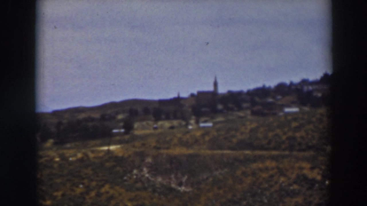 1937 Truckee countryside frame showing brushy slopes, small distant structures, and a steeple on the horizon line.
