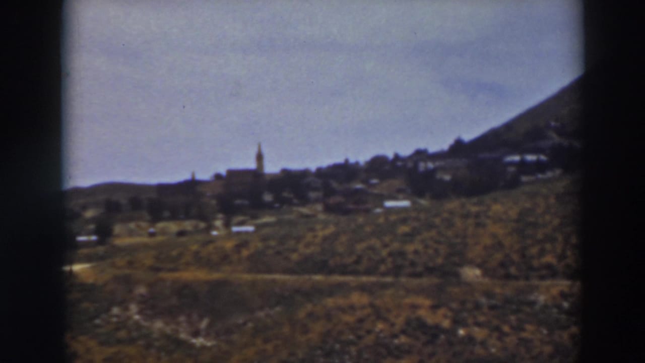Distant hillside town with a steeple, low buildings, scrub foreground, and pale sky.