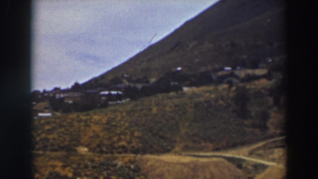 Dirt road and scattered buildings on a brushy hillside beneath a mountain.
