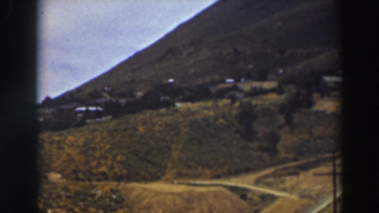 1937 Truckee frame showing brushy fields, a curving dirt road, distant buildings, and a steep mountain slope in soft 8mm color.