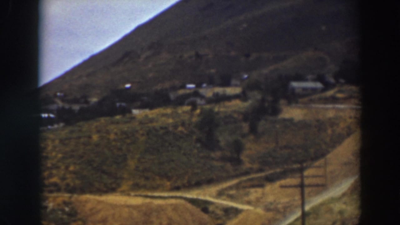 1937 Truckee home-movie frame with a fence post in the foreground, a dirt road, dry fields, and distant hillside buildings.