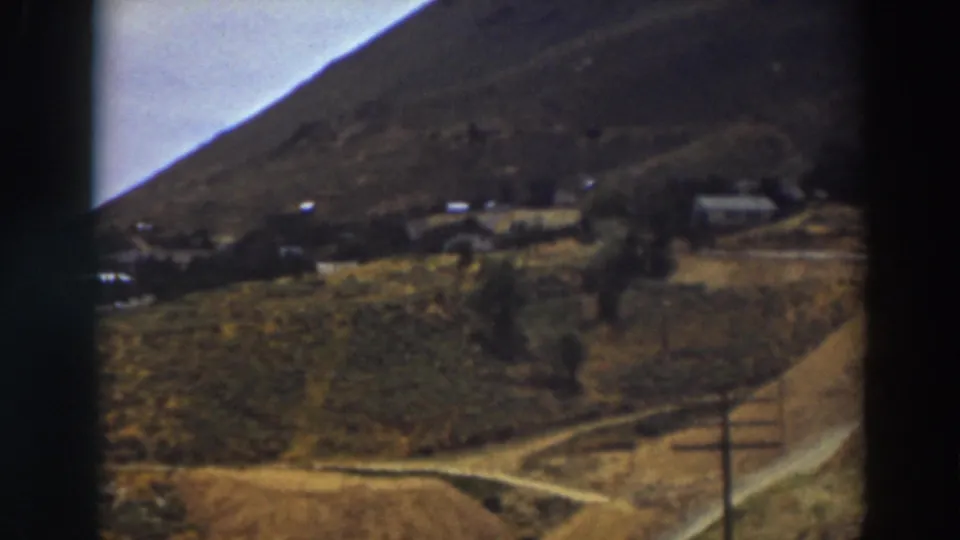 1937 Truckee home-movie frame with a fence post in the foreground, a dirt road, dry fields, and distant hillside buildings.