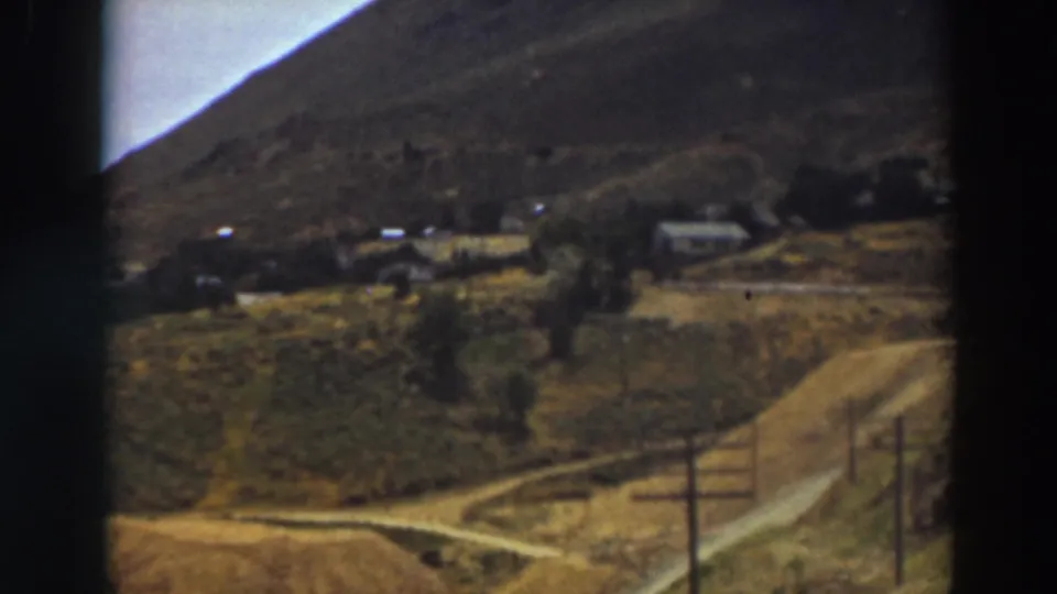 Truckee-area mountain valley view on 8mm film, with small buildings, a dirt road, utility poles, and open land under a hillside.