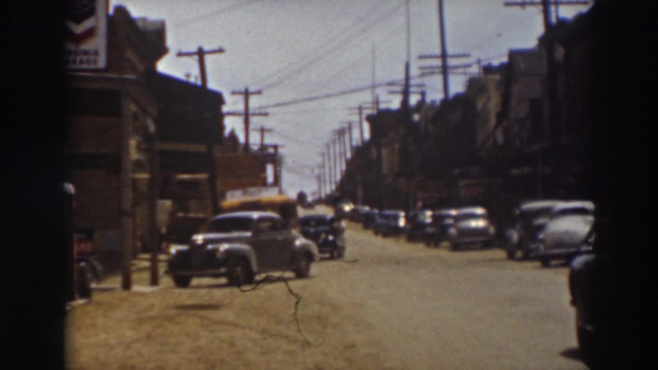 Late-1930s cars sit along a Truckee street beneath overhead wires, with storefronts at left and right and a partial dark figure at the edge.