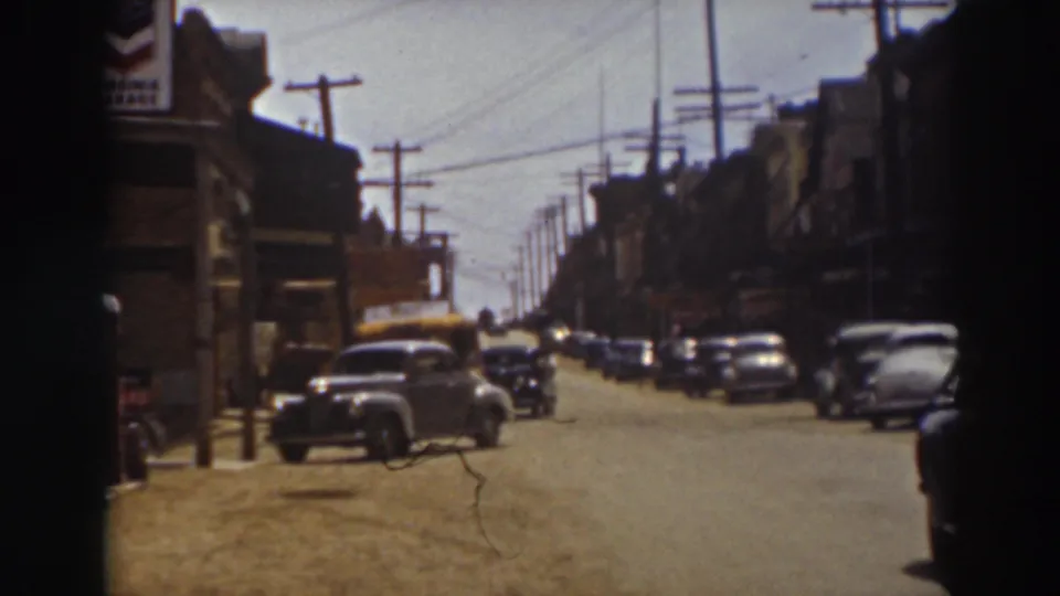 Late-1930s cars sit along a Truckee street beneath overhead wires, with storefronts at left and right and a partial dark figure at the edge.