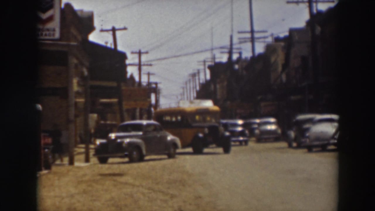 1937 Truckee footage showing a yellow bus moving past a row of parked cars, with storefronts and utility wires framing the street.