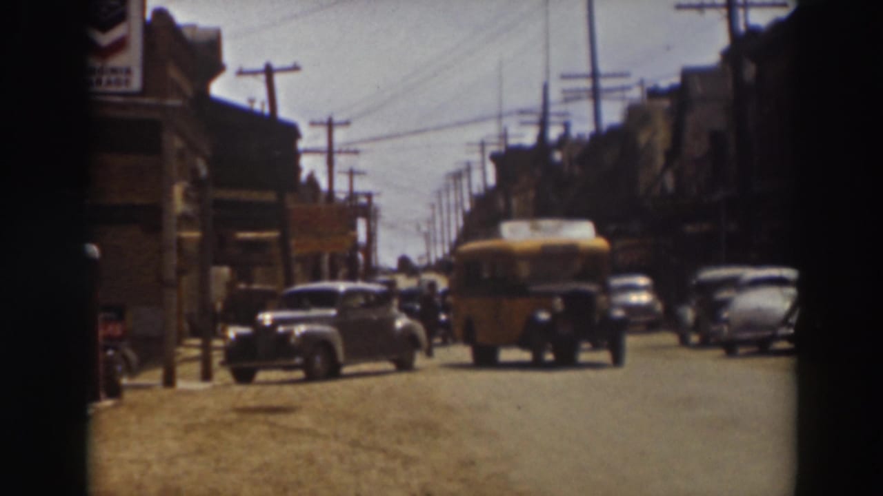 Yellow school bus approaches on a dusty street with parked cars and storefronts.