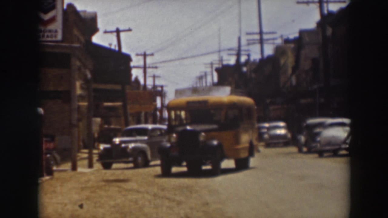 Nearer view of a yellow school bus traveling down the Truckee street, with a gray sedan, parked cars, buildings, and overhead wires around it.