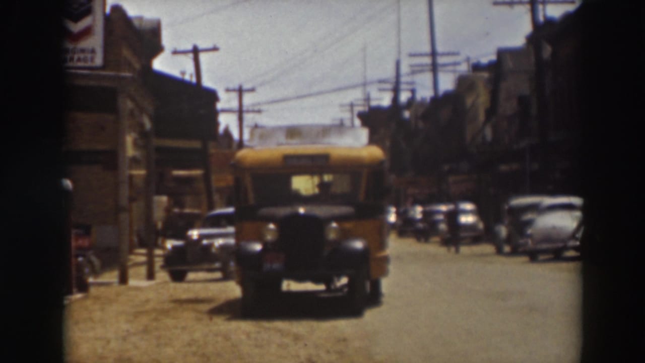 The front of the yellow school bus reaches the foreground on a 1937 Truckee street, framed by parked cars, storefronts, poles, and wires.