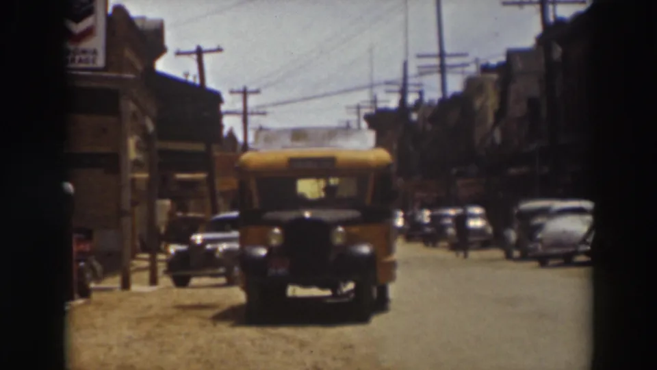 The front of the yellow school bus reaches the foreground on a 1937 Truckee street, framed by parked cars, storefronts, poles, and wires.