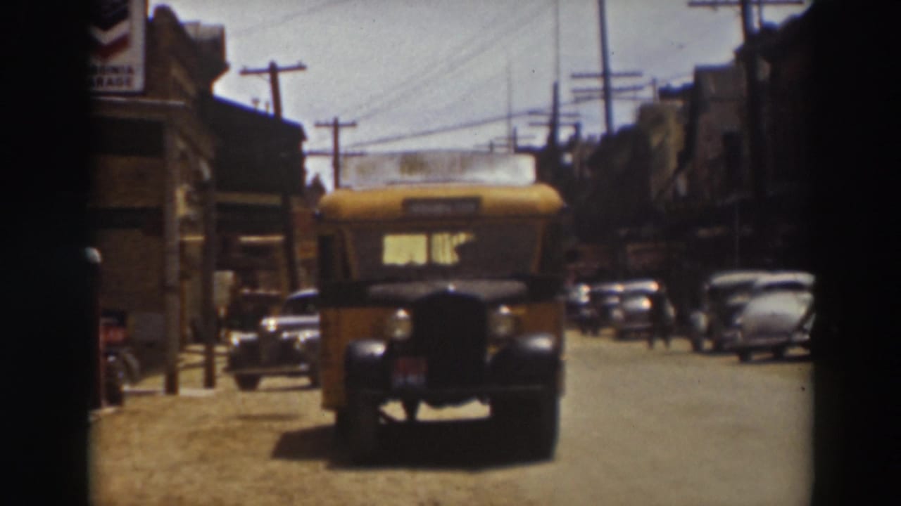 Yellow bus driving toward the camera on a sunlit street with parked cars and utility poles.