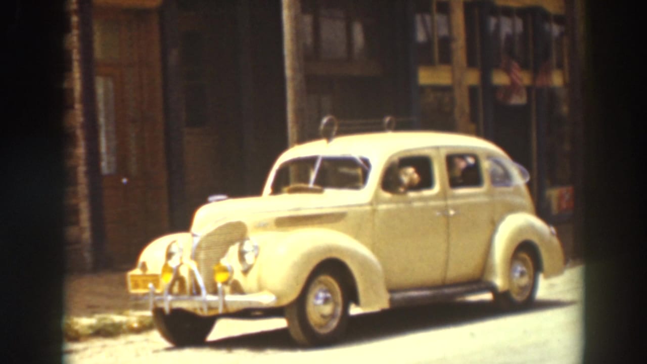 Cream-colored 1930s sedan parked beside Truckee storefronts, with a visible occupant and bright sunlight on the rounded fenders.
