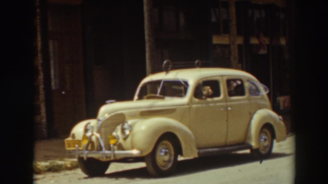 Truckee street view of a cream 1930s sedan at the curb, with visible occupants, storefront shadows, and the car's rounded bodywork.