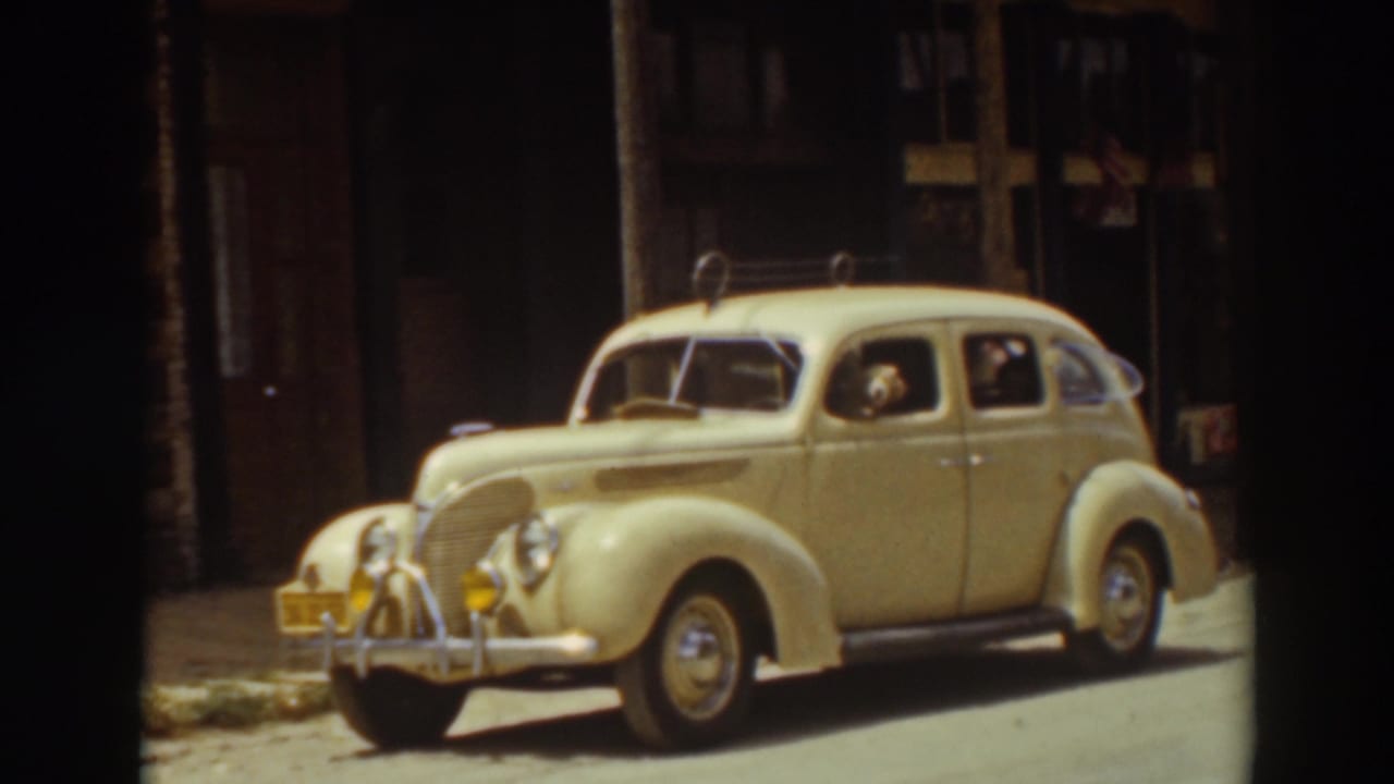1937 Truckee home-movie frame showing a cream-yellow sedan parked on a sunny street, with a dog and people visible through the windows.