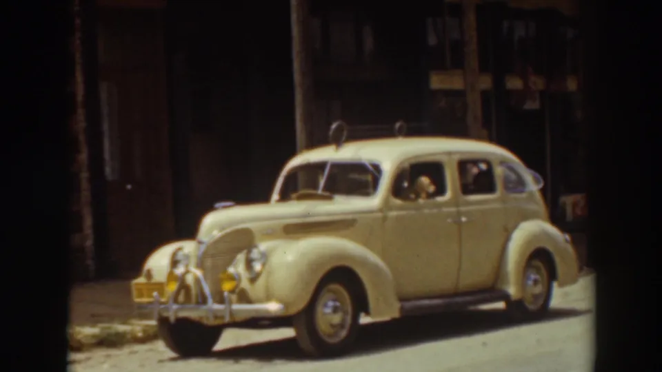 1937 Truckee frame of a cream-colored classic sedan beside a dark storefront, with a dog peering from the passenger-side window.