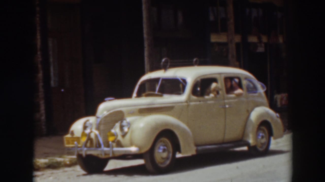 Cream-colored vintage sedan on a quiet street with a driver and dog visible through the side windows.