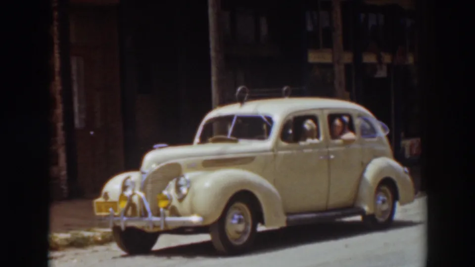 1937 Truckee frame of a rounded cream sedan moving along the street, with the dog still visible at the passenger window and the driver behind the wheel.