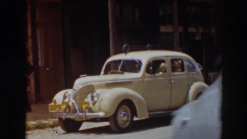 A passing vehicle crosses the foreground while the cream-colored 1937 street sedan remains parked near buildings in Truckee.