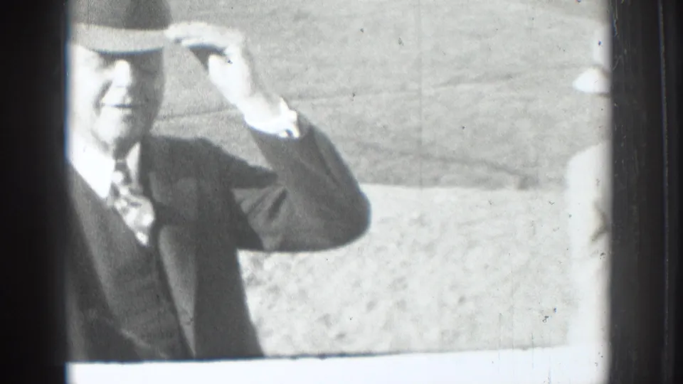 Black-and-white 8mm frame of an adult man in a suit and brimmed hat raising his hand near the hat in Vacaville, California, 1937.