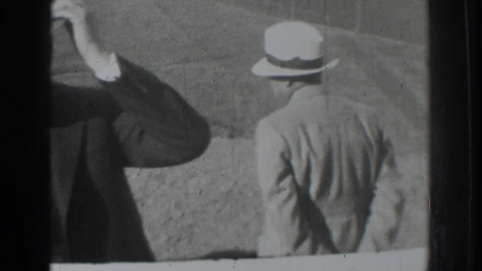 Black-and-white 8mm frame shows a cropped suited man raising a hand beside another hat-wearing man seen from behind in Vacaville, 1937.