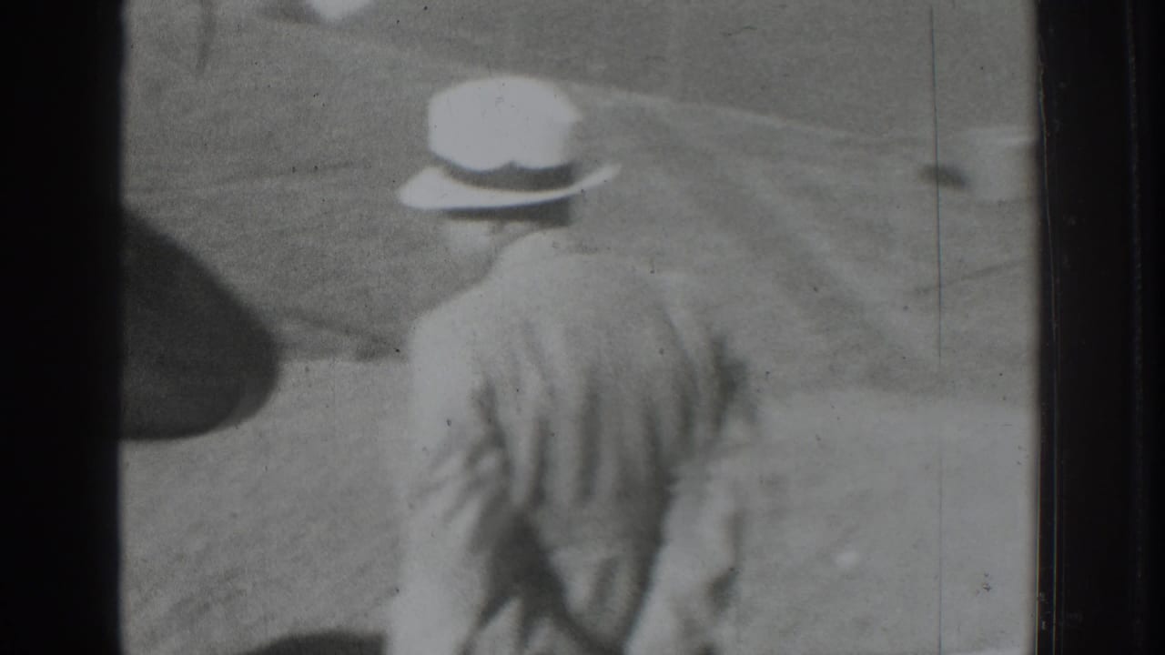 Man in a light suit and white brimmed hat walking outdoors near low hills.