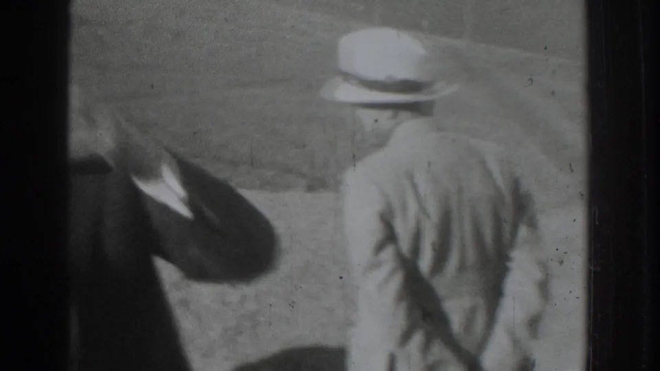 A man in a dark suit appears to look through binoculars as a light-suited man walks ahead in the Vacaville hillside setting.