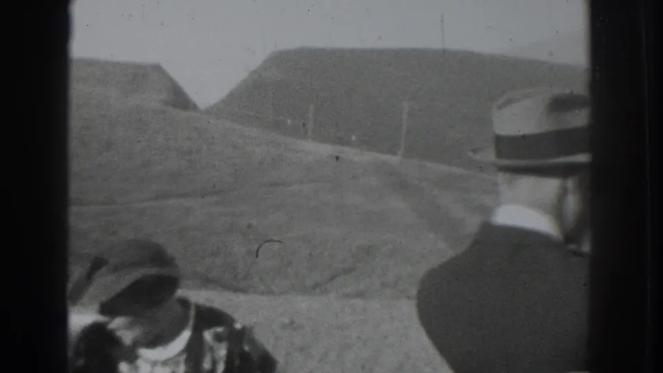 1937 Vacaville home-movie frame with two adult men in hats near a vehicle, backed by rolling hills and a dirt road.