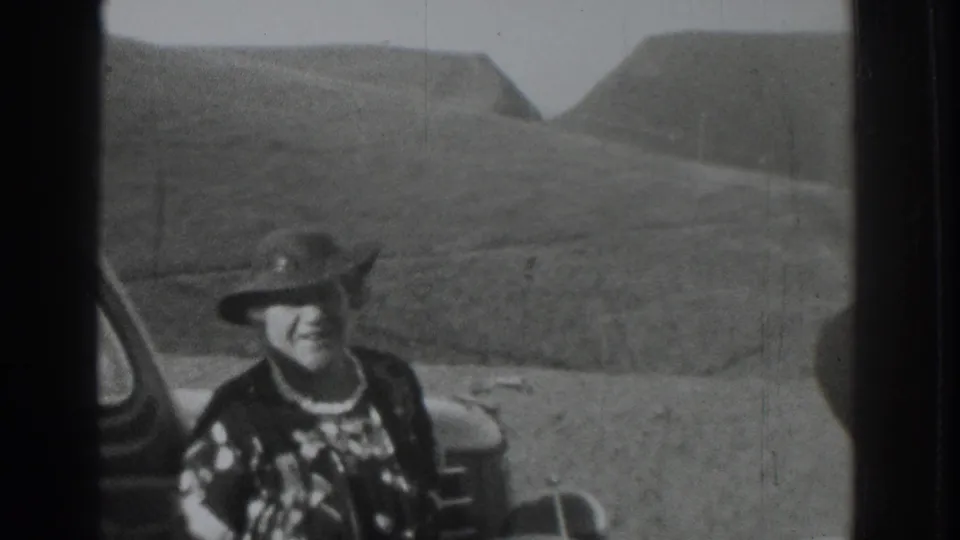 In supplied 1937 Vacaville footage, a woman in a hat and patterned dress smiles near a car at a rural hillside overlook.