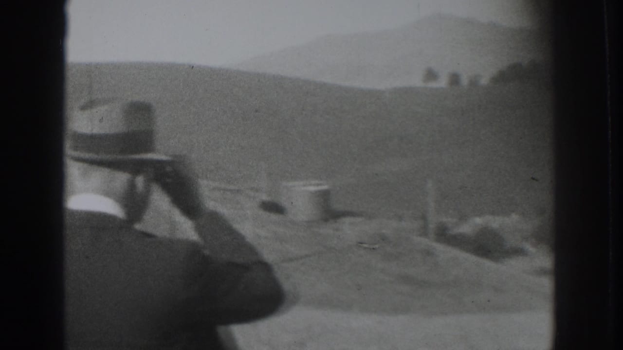 1937 Vacaville home-movie frame showing a suited man in a fedora looking through binoculars across a rural hillside.